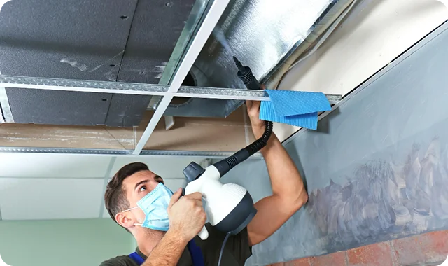 A man wearing a face mask uses a sprayer on a ceiling, installing insulation in an unfinished room. The setting conveys a sense of concentration and technical work.