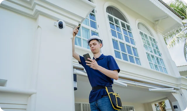 A technician in a blue shirt and helmet is installing a security camera on a white home's exterior. He holds a smartphone, appearing focused and professional.