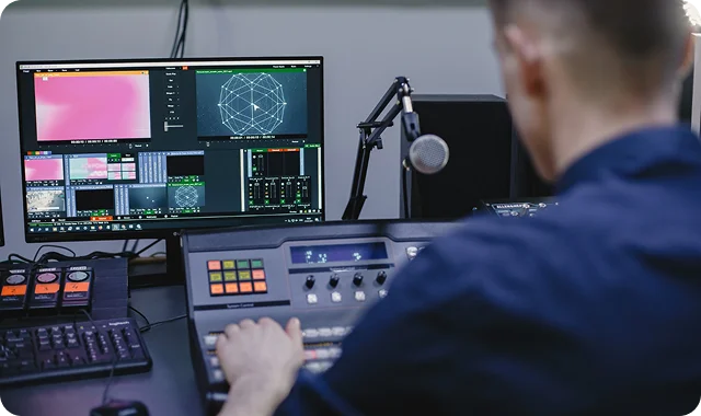 A person operates an audio mixing console in a studio, with multiple monitors displaying graphics and controls. The scene conveys focus and professionalism.