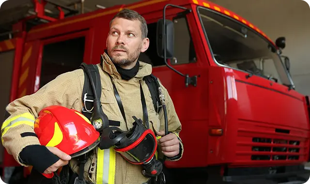 Firefighter in full gear holds a helmet and mask, standing confidently beside a red fire truck.
