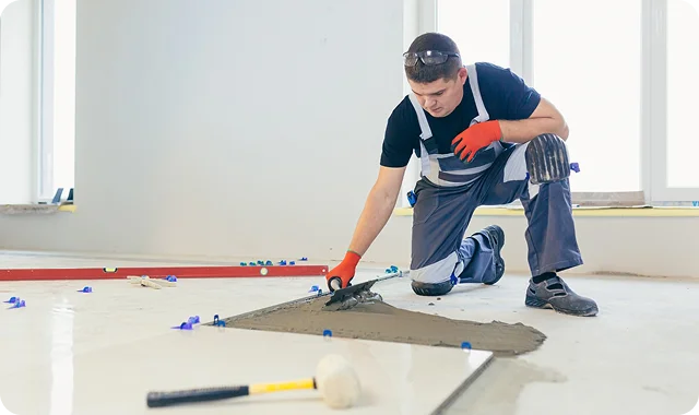 A construction worker in gloves and kneepads is kneeling, smoothing concrete on a floor with a trowel. The scene conveys focus and precision.