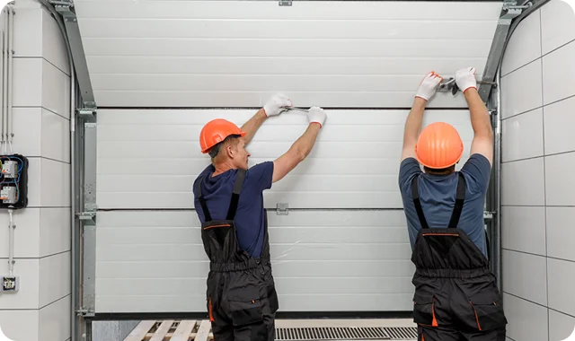 Two workers in orange hard hats and gloves install a garage door, focused on ensuring it fits correctly within the frame.