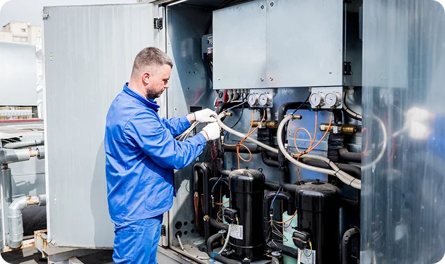 A technician in blue overalls and gloves inspects industrial HVAC equipment on a rooftop, adjusting gauges with focused attention.